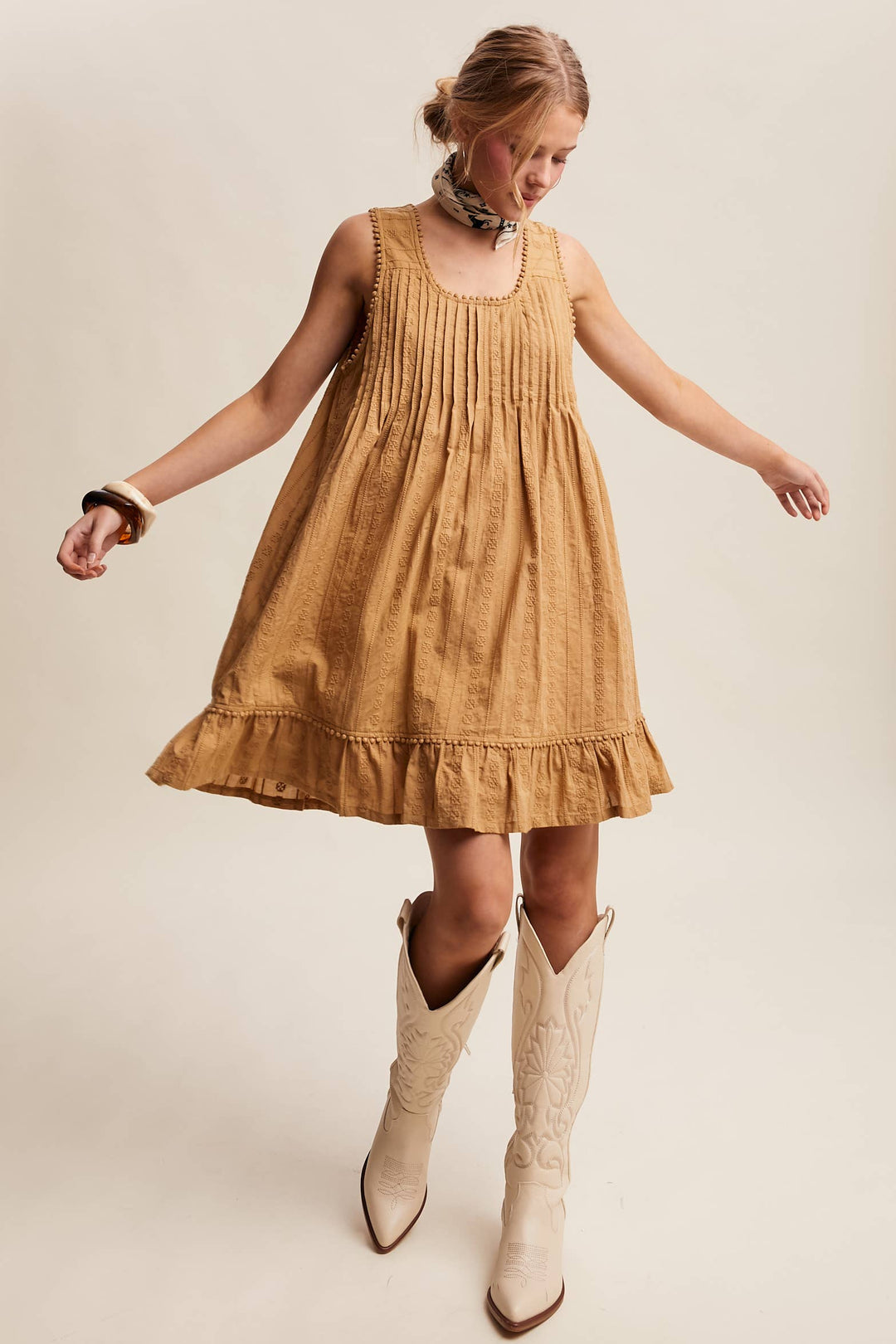 Woman wearing a textured camel-colored sleeveless mini dress with pintuck details and ruffle hem.
