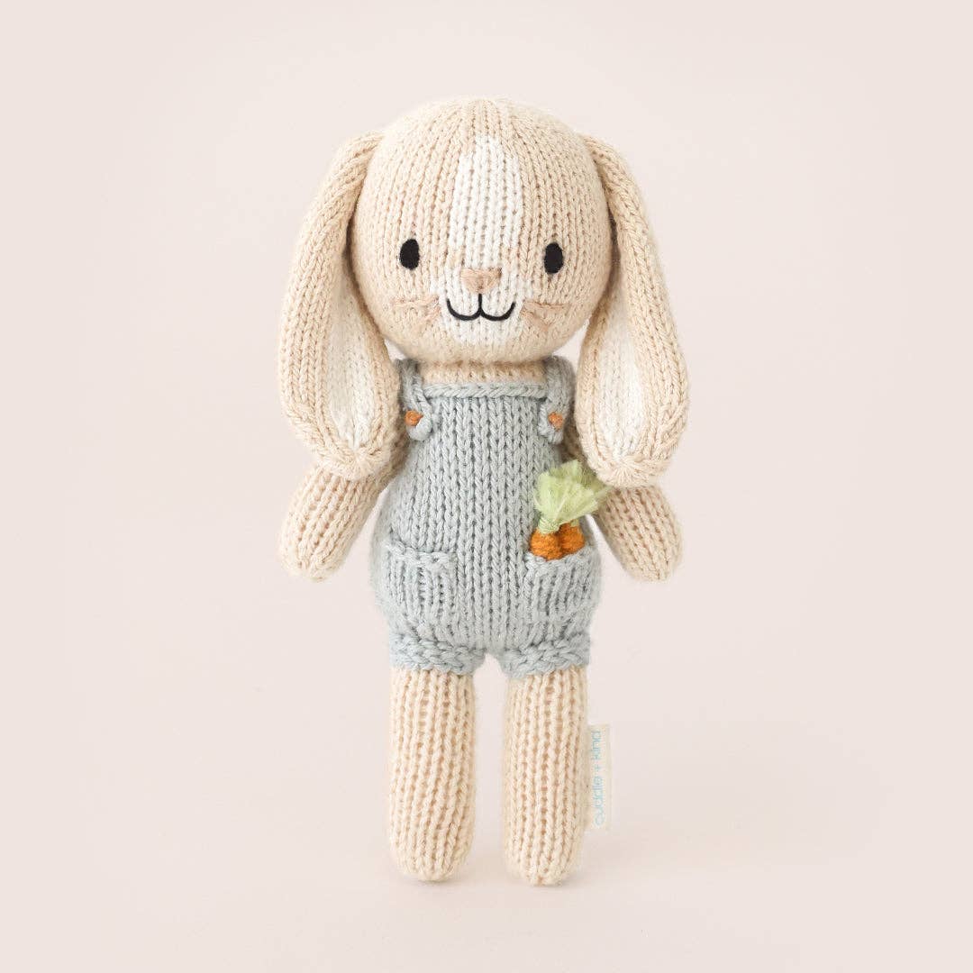 Knitted toy rabbit in overalls holding a carrot on a light beige background
