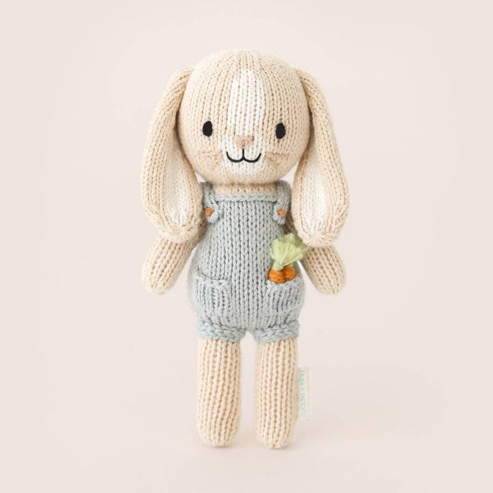 Knitted toy rabbit in overalls holding a carrot on a light beige background