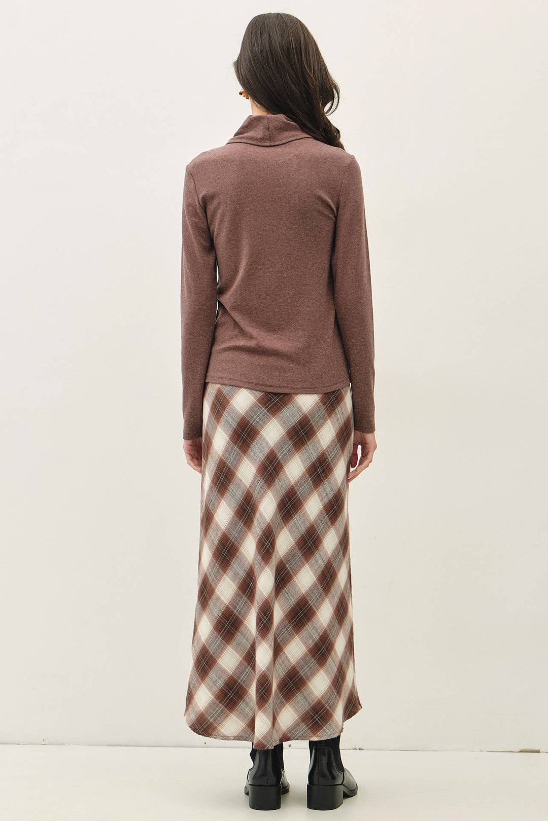 A woman viewed from behind wearing a brown long-sleeve turtle-neck top and a brown, beige, and white plaid maxi skirt, paired with black boots, against a plain off-white studio background.