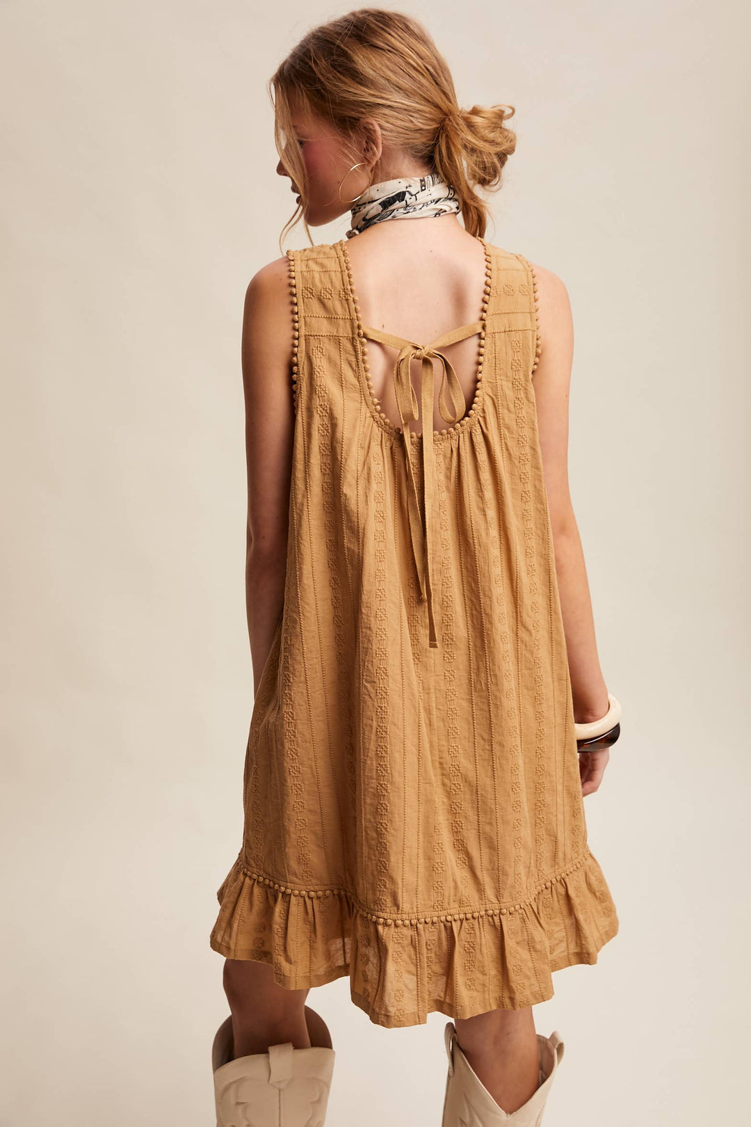 Woman wearing a textured camel-colored sleeveless mini dress with pintuck details and ruffle hem. Shows back and bow/tie detail