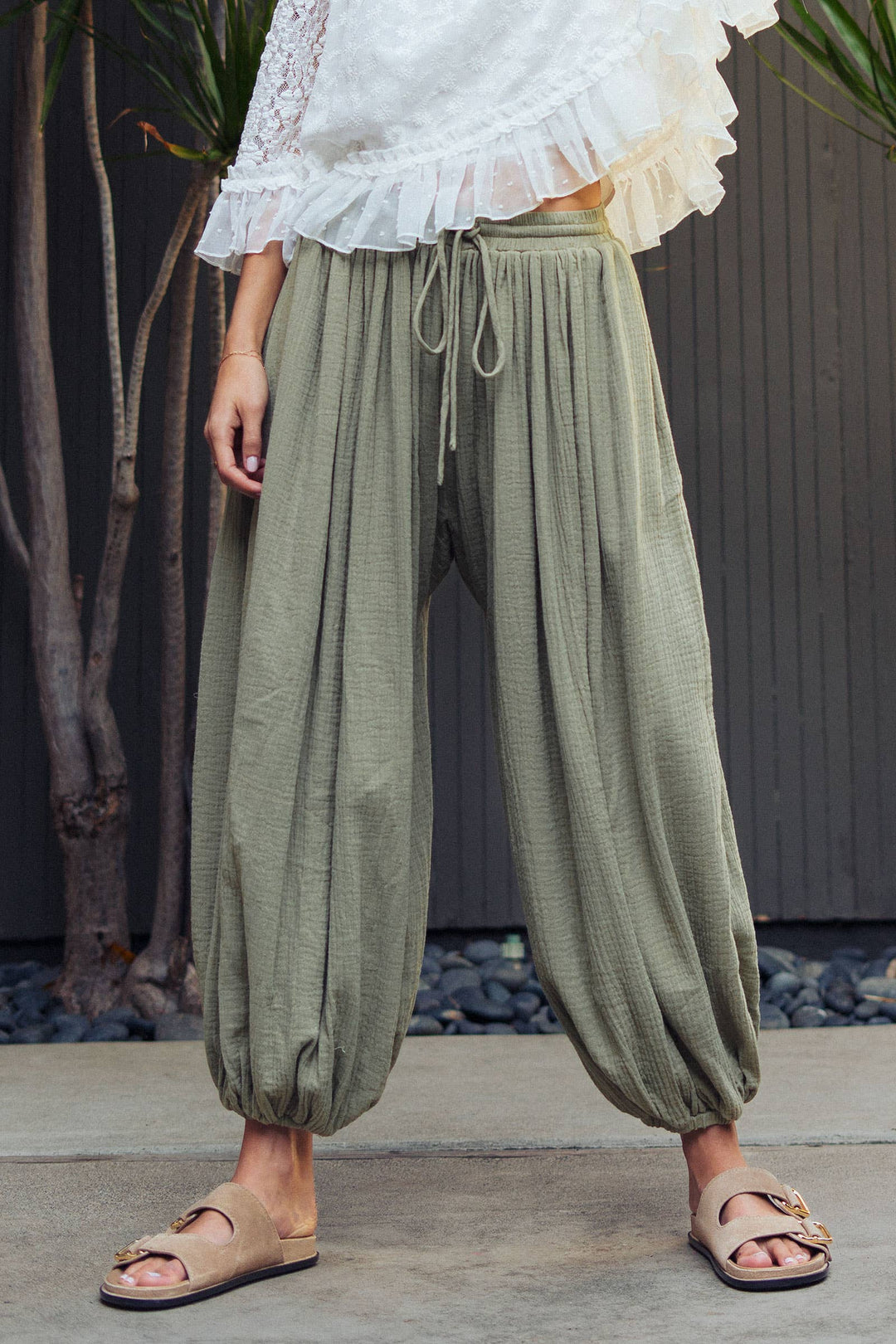 Woman wearing olive green crinkle cotton balloon pants with drawstring waist and elastic ankle cuffs paired with a white lace blouse.