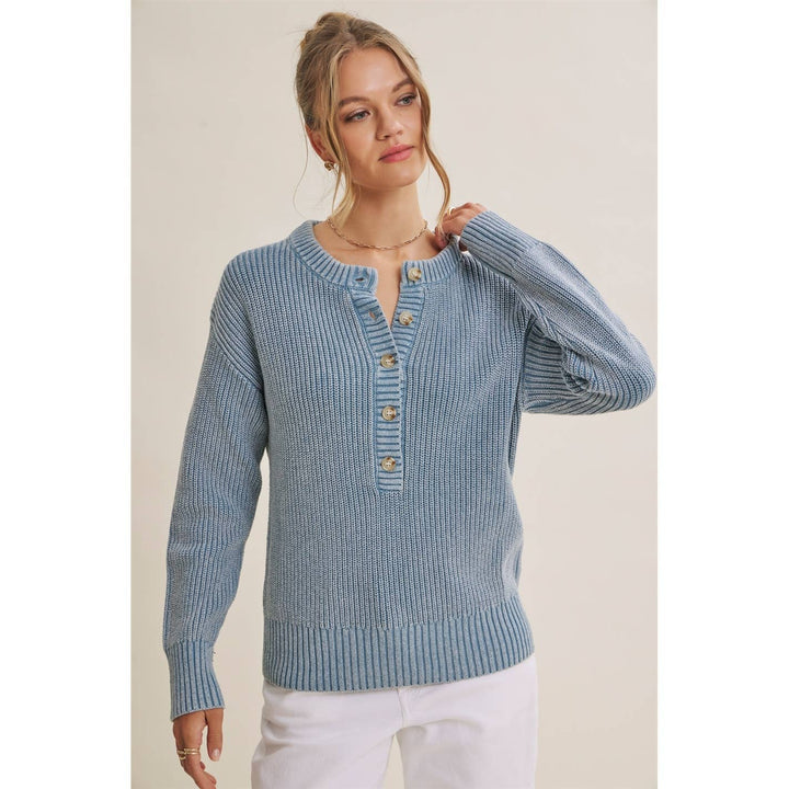 A woman wearing a light blue ribbed knit sweater with a buttoned henley neckline and white pants, standing against a plain beige studio background.