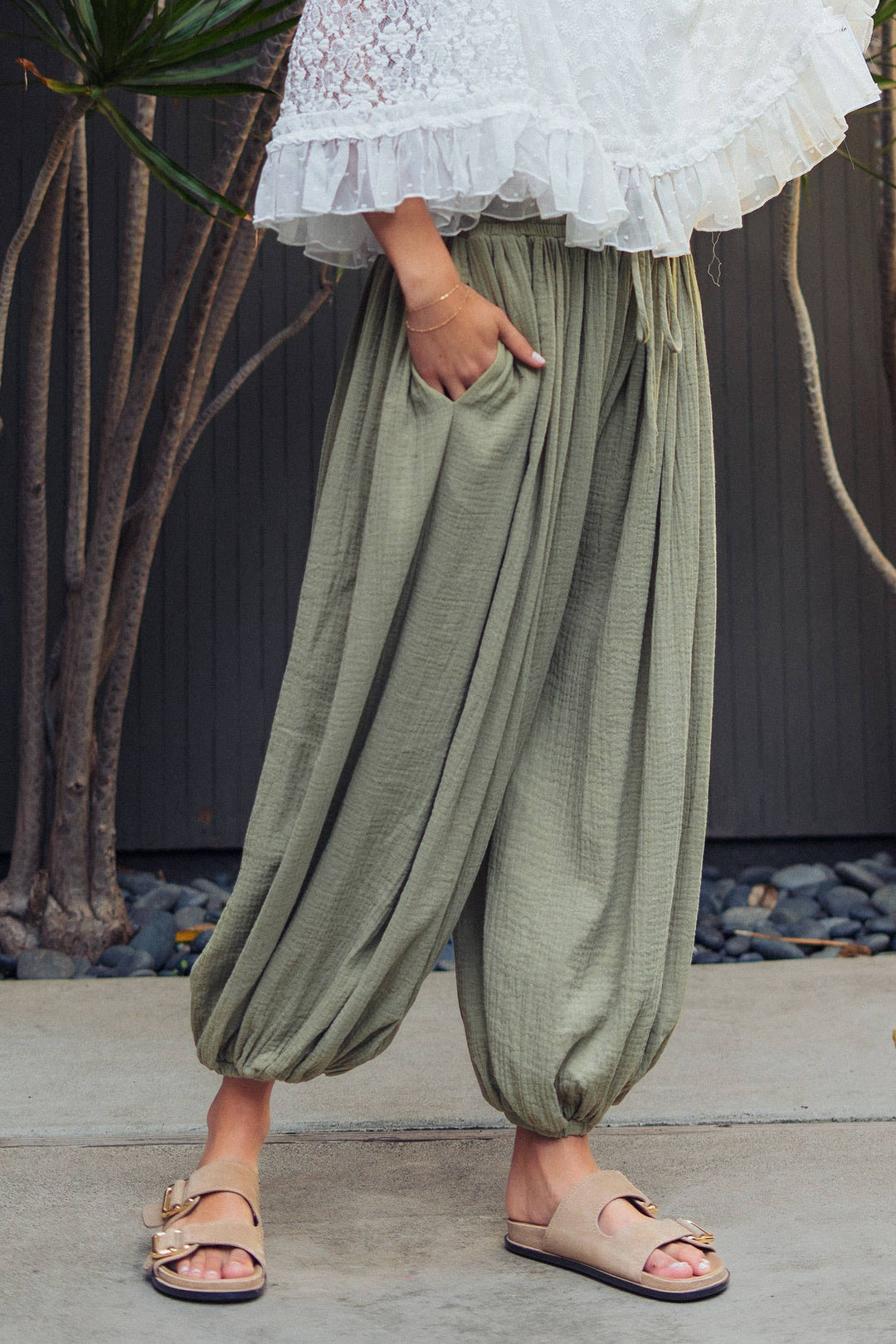Woman wearing olive green crinkle cotton balloon pants with drawstring waist and elastic ankle cuffs paired with a white lace blouse.
