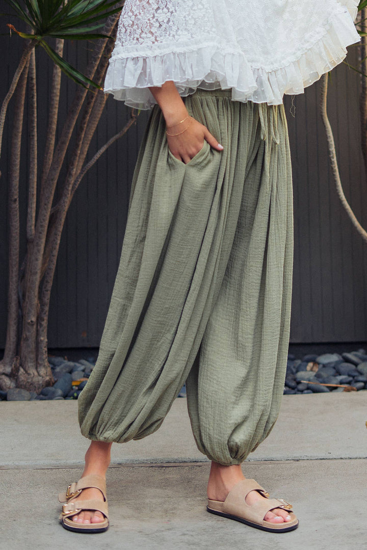 Woman wearing olive green crinkle cotton balloon pants with drawstring waist and elastic ankle cuffs paired with a white lace blouse.