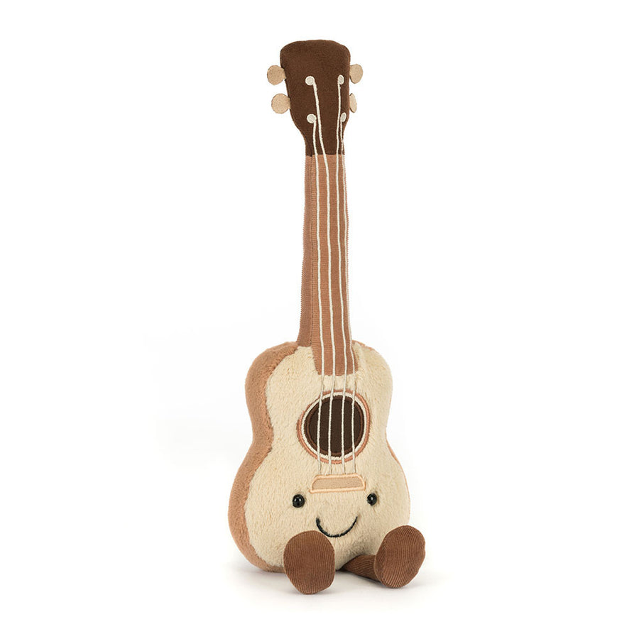 A soft, plush Jellycat Amuseables Ukulele toy with a light beige body, dark brown neck and headstock, black eyes and a smiling mouth, a brown soundhole, and white 'strings,' sitting with two corduroy brown feet extended forward on a white background.