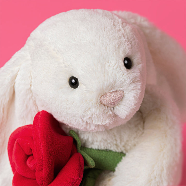 closeup detail shot Jellycat Cream Bunny plush holding a red rose with soft cream fur and floppy ears on a pink background