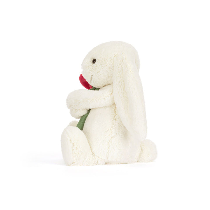 Jellycat Cream Bunny plush holding a red rose with soft cream fur and floppy ears on a white background left facing view