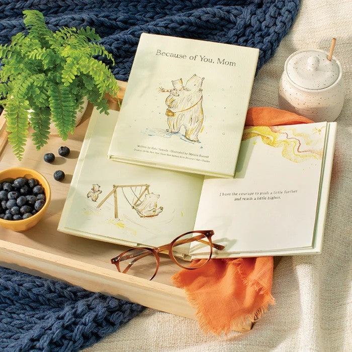 Book titled 'Because of You, Mom' on a wooden tray with glasses, fruit, and a plant.