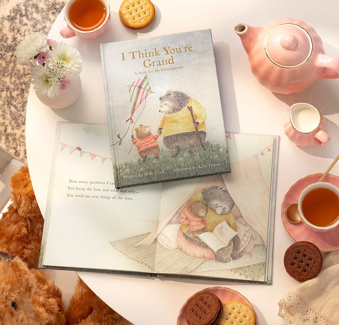 Book titled 'I Think You're Grand' on a table with tea, cookies, and a teddy bear.