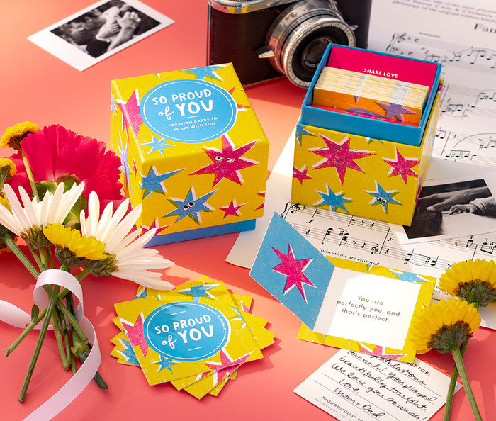 Colorful 'So Proud of You' card set with flowers and a camera on a pink surface