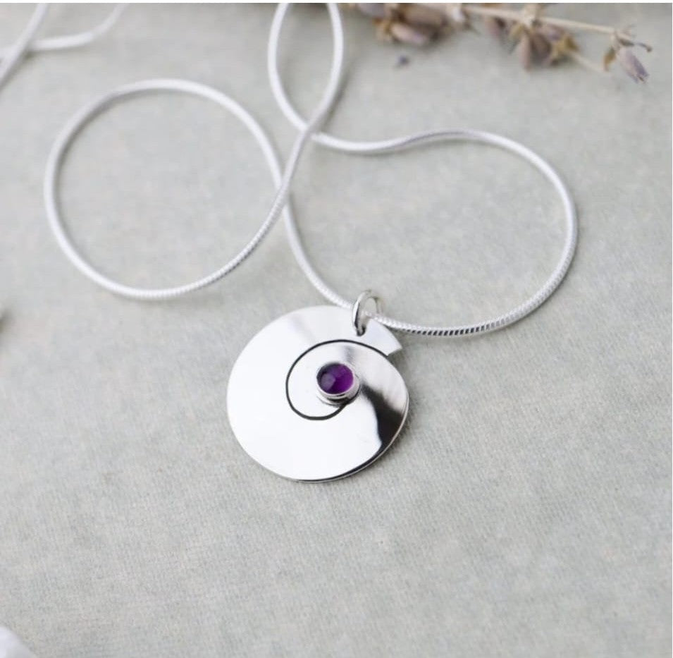 Silver necklace with a circular pendant featuring an amythest gemstone on a light gray background.