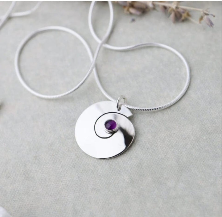 Silver necklace with a circular pendant featuring an amythest gemstone on a light gray background.
