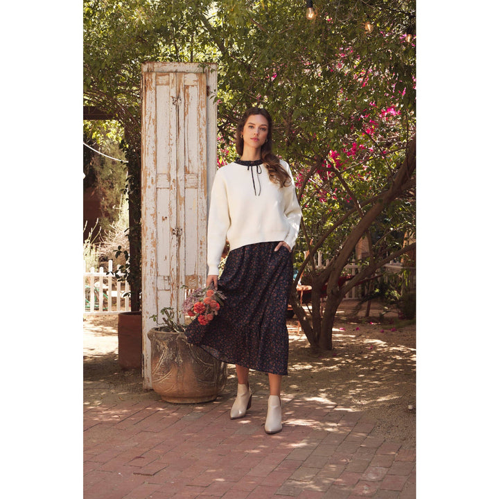Model wearing Bow Back Sweater | Dolman Sleeve Knit with Satin Tie. Lifestyle photo with outdoor garden setting.  Distance shot showing sweater layered with a black blouse paired with floral skirt & neutral booties.