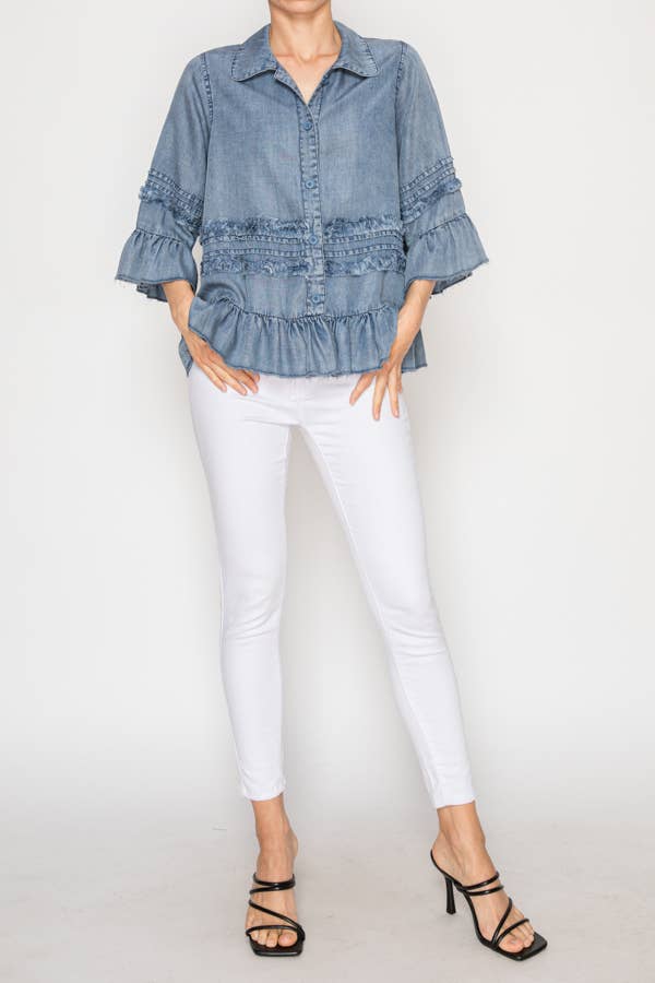 A model is shown from the waist up wearing a light blue chambray button-down top with horizontal ruffle details and flared three-quarter sleeves. She has her hands in the pockets of her white skinny jeans and is wearing black strappy heels.

