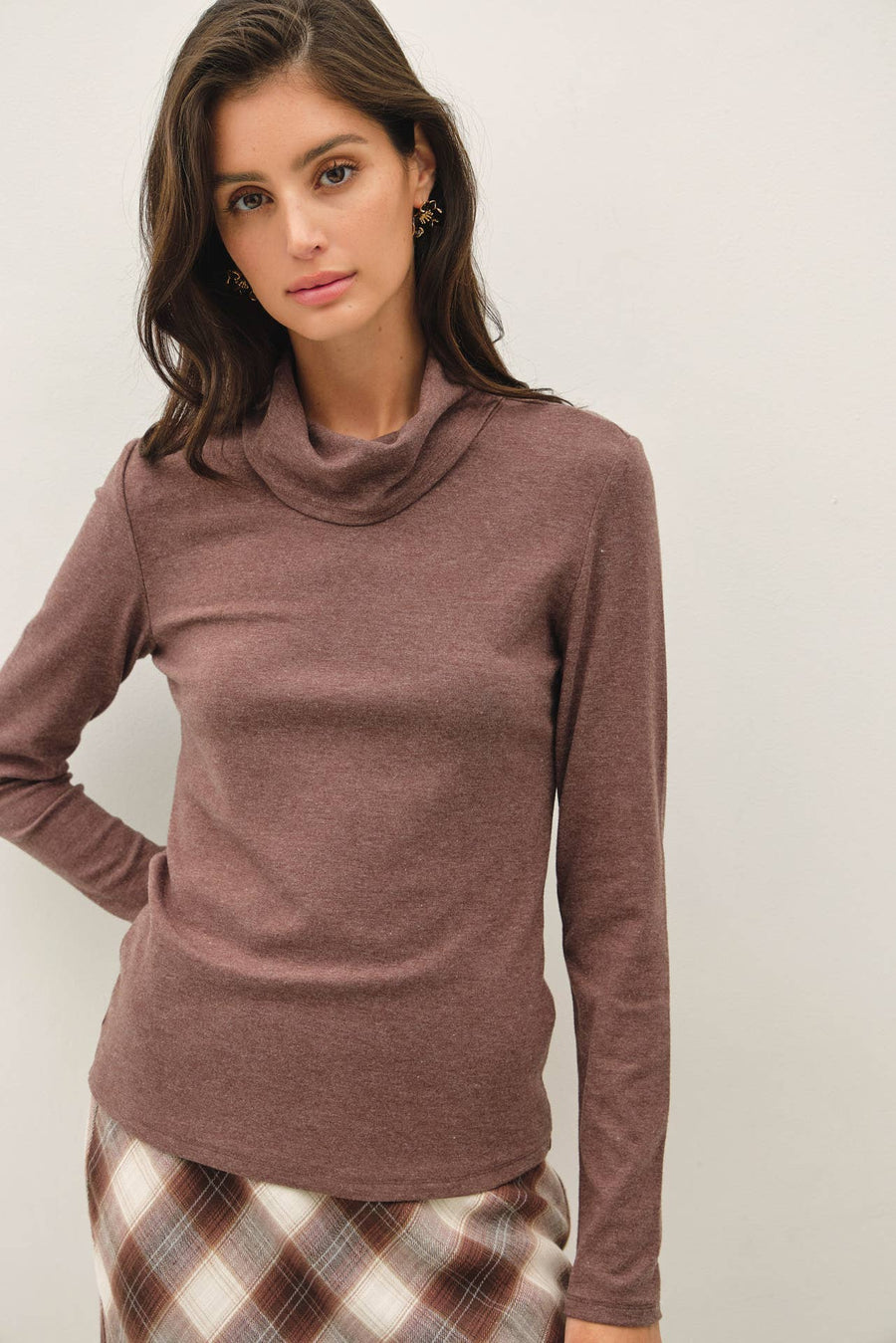 A woman wearing a long-sleeve turtle neck knit top in a dusty mauve-brown color, paired with a brown and white plaid skirt, against a plain light gray background.