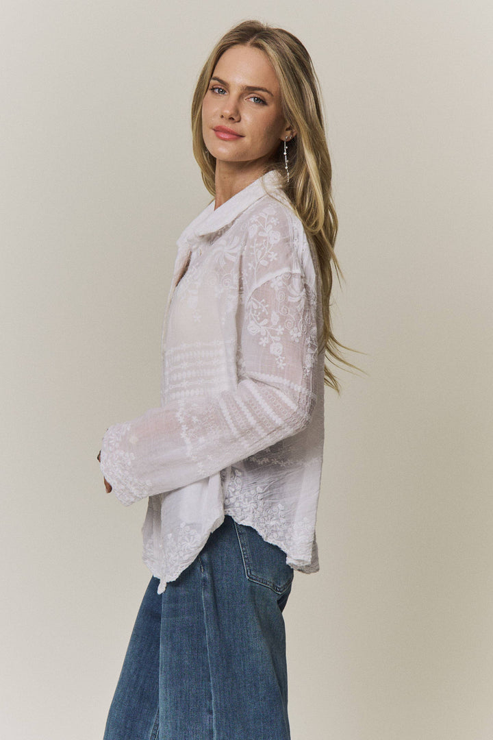 Woman wearing a light-colored lace embroidered blouse and blue jeans against a plain background
