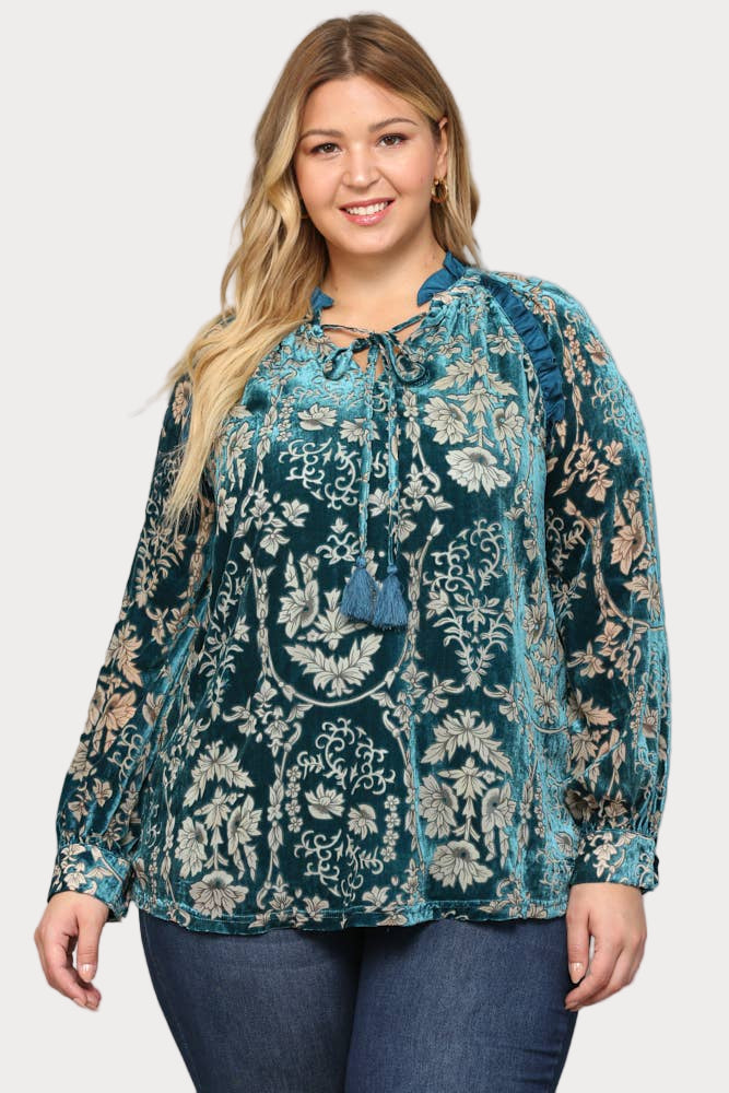 A smiling plus-size model wears a long-sleeve teal burnout velvet blouse featuring a floral pattern, ruffle trim, and a tassel tie at the neck.

