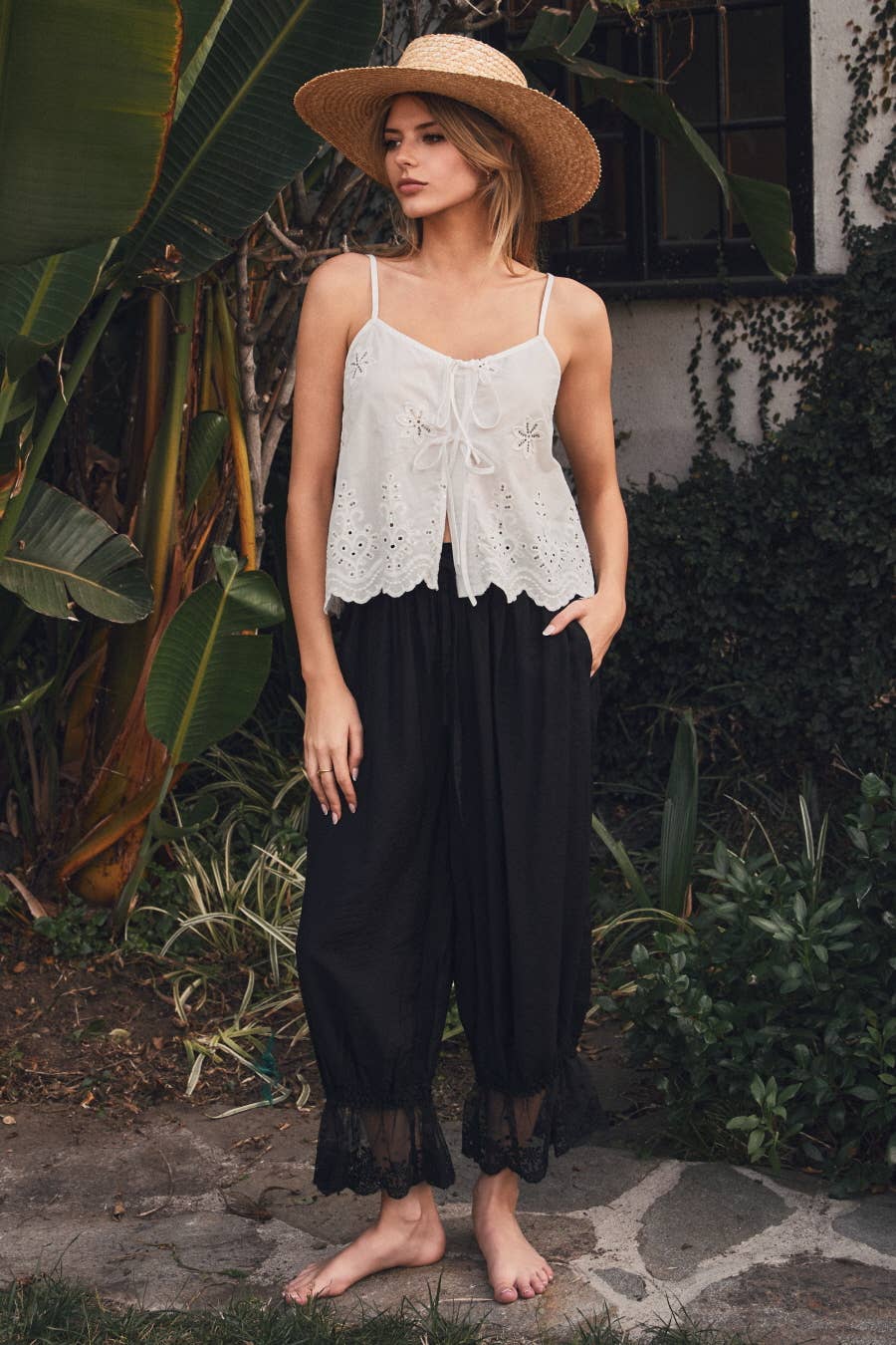 Woman wearing black relaxed lace hem pants with an elastic drawstring waistband and scalloped lace cuffs paired with a white embroidered camisole.
