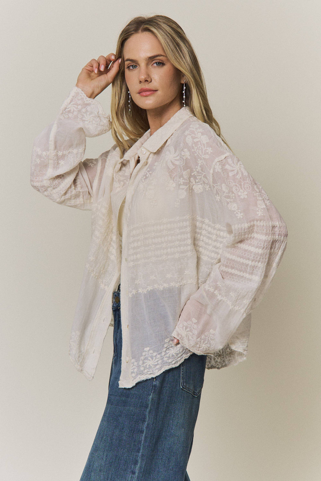 Woman wearing a light-colored lace embroidered blouse and blue jeans against a plain background