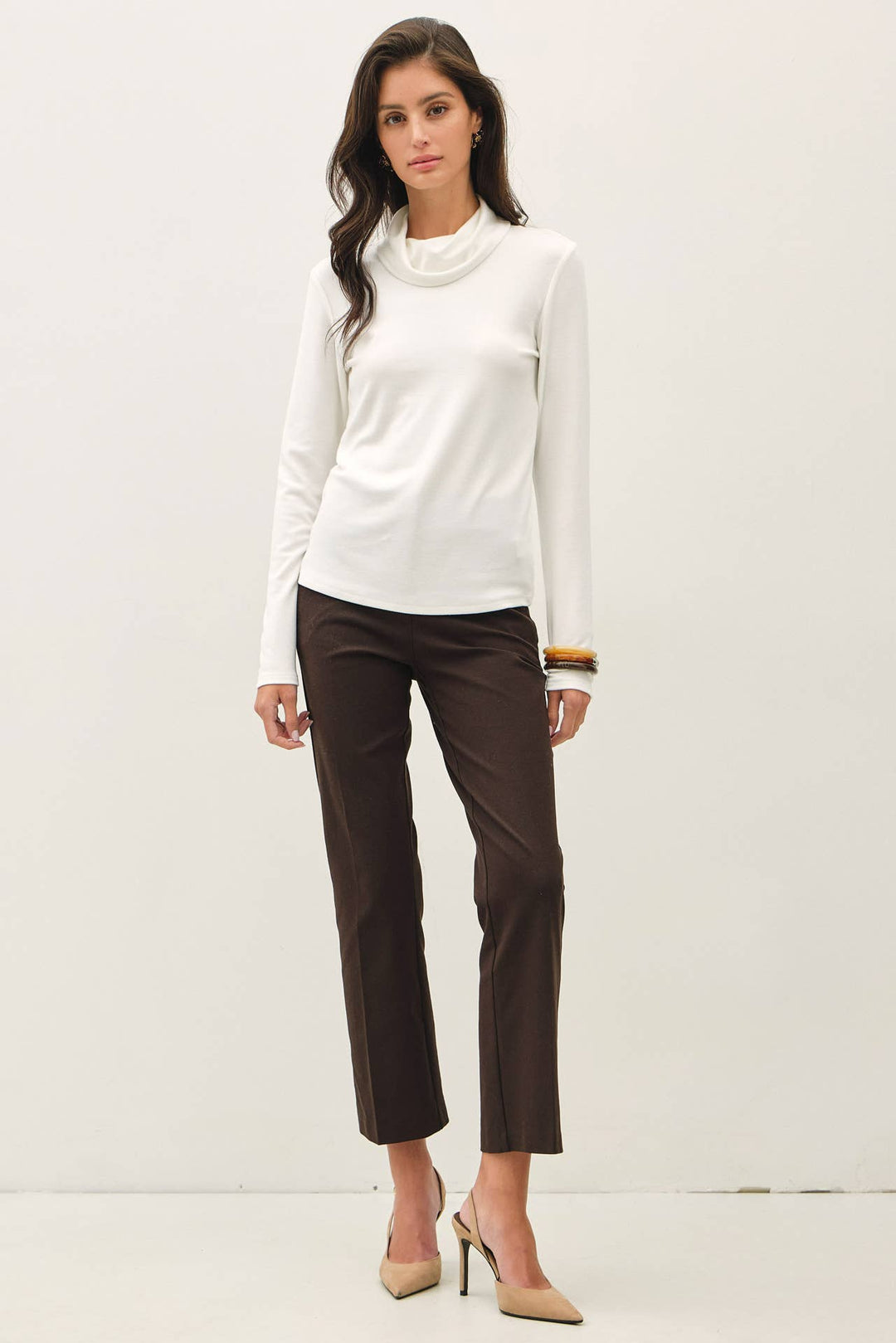 A woman modeling a white long-sleeve turtle neck top and dark brown cropped trousers with beige heels, standing against a plain white studio background.