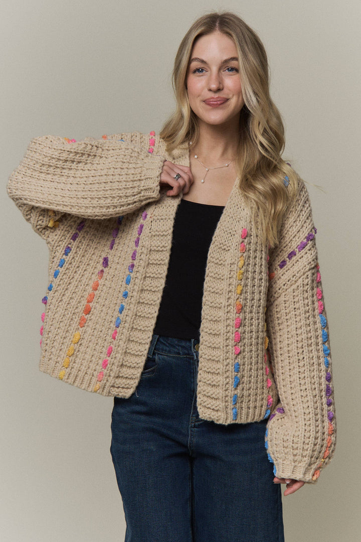 A woman wearing a chunky knit cardigan with a beige base and colorful vertical stripe patterns in pink, blue, yellow, and purple, styled with a black top and blue jeans against a solid beige background.