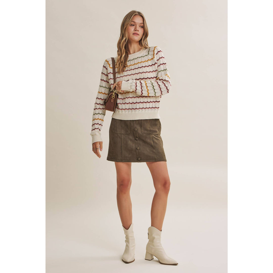 A woman modeling a cream knit sweater with multicolor horizontal stripes, paired with an olive green mini skirt and cream ankle boots, against a solid light beige background.