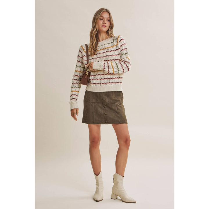A woman modeling a cream knit sweater with multicolor horizontal stripes, paired with an olive green mini skirt and cream ankle boots, against a solid light beige background.