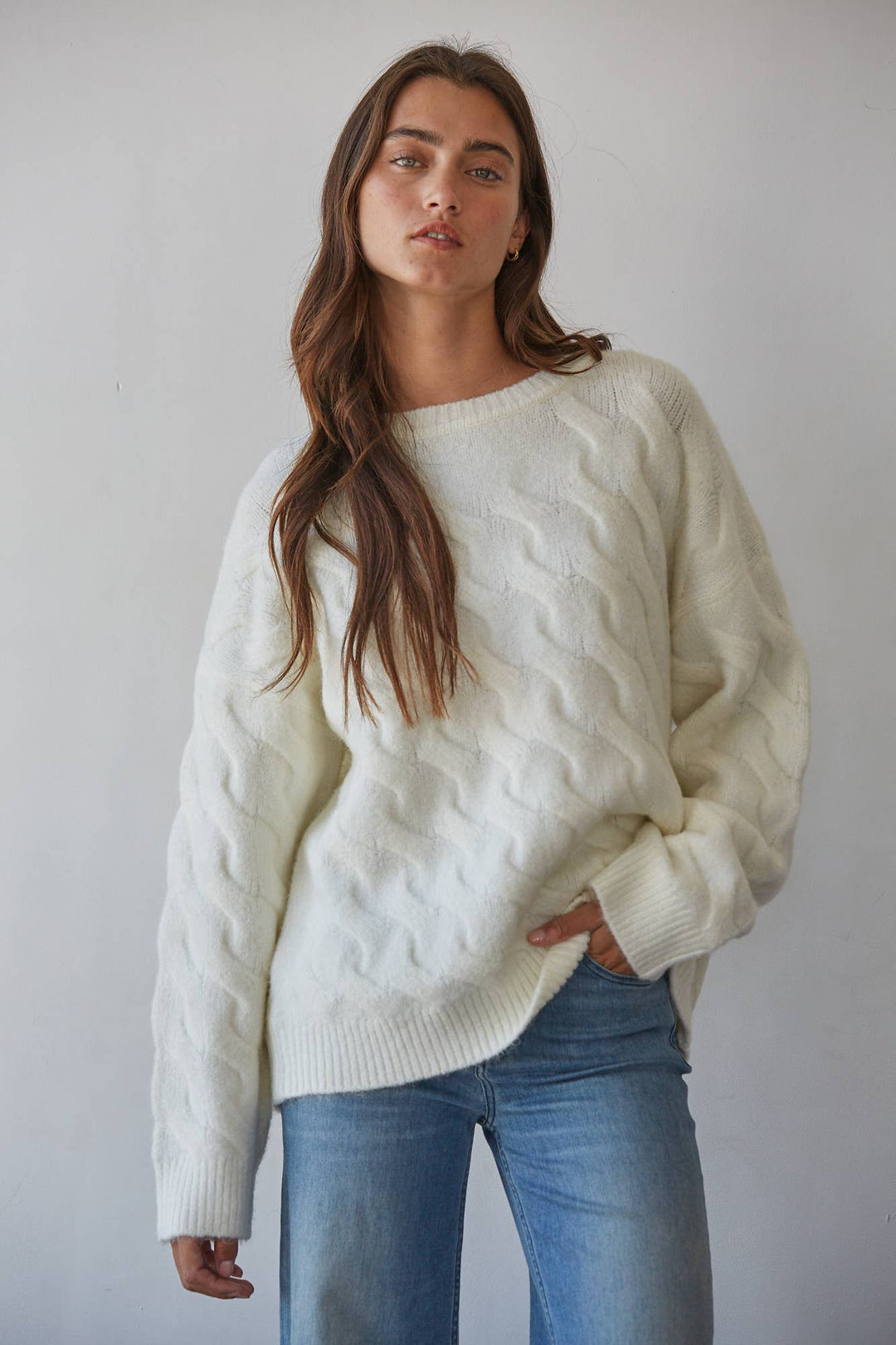 A woman wearing an oversized cream cable-knit sweater and blue jeans, standing against a plain light gray wall.