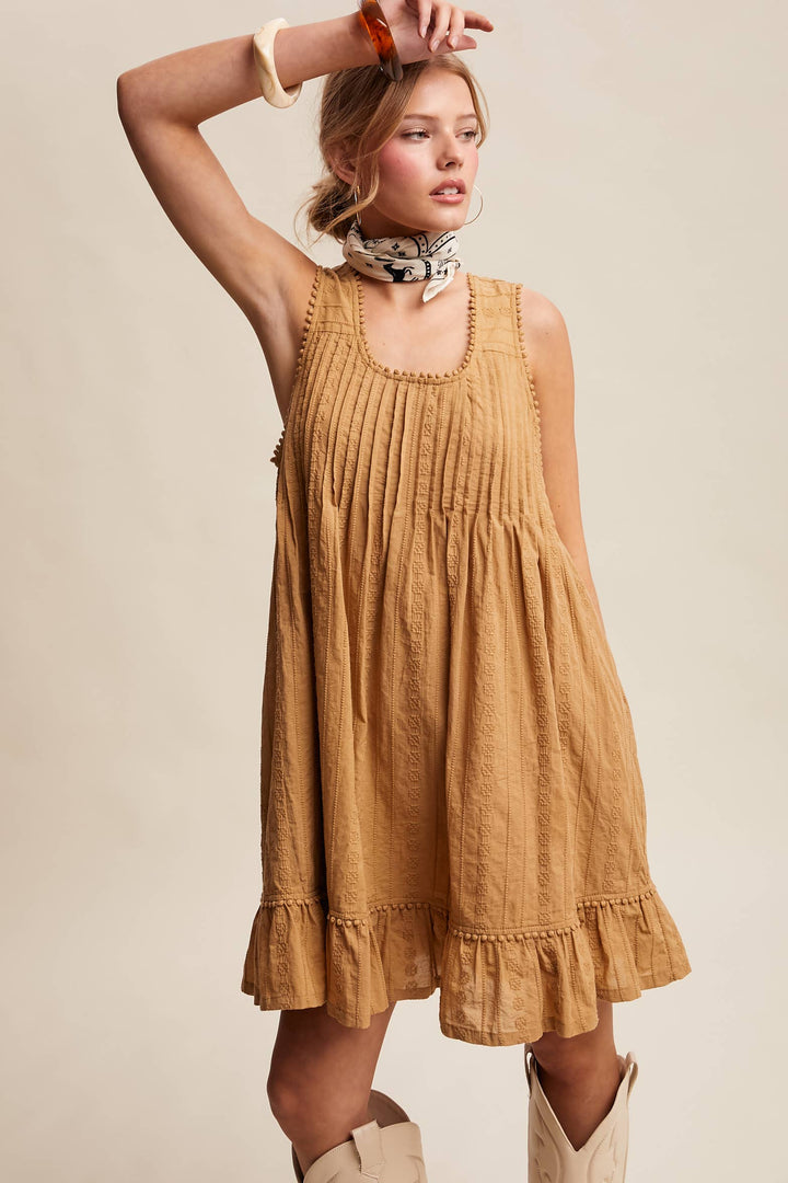 Woman wearing a textured camel-colored sleeveless mini dress with pintuck details and ruffle hem.