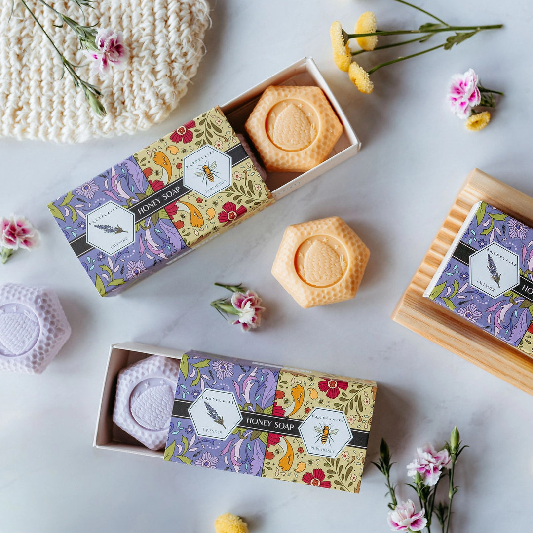 Three hexagonal honey soap bars arranged with their decorative packaging on a light gray surface, styled with small flowers and a knitted textile.