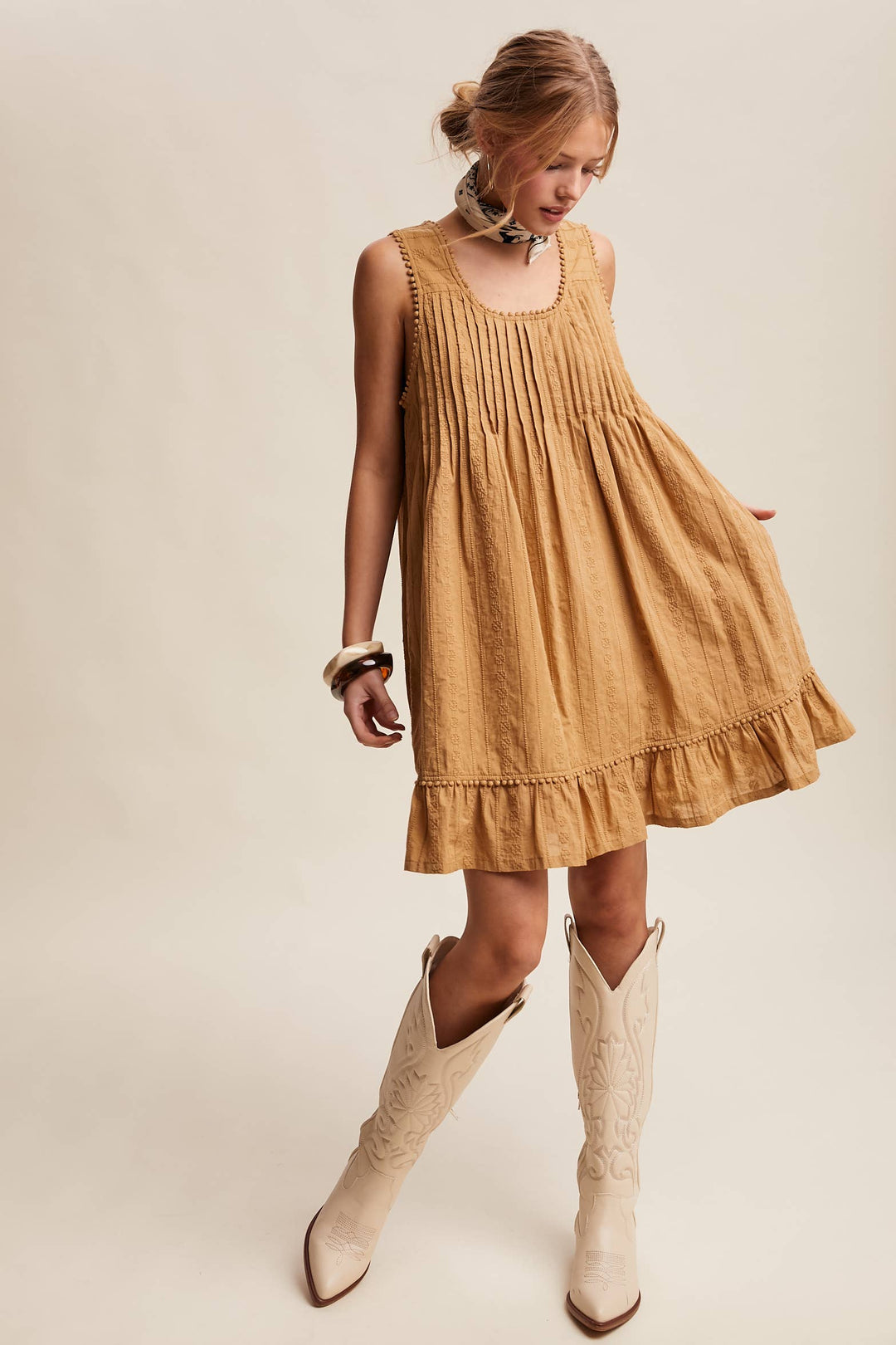 Woman wearing a textured camel-colored sleeveless mini dress with pintuck details and ruffle hem.