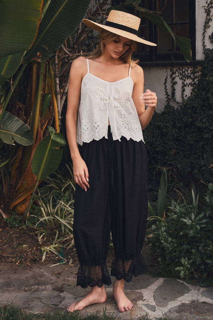 Woman wearing black relaxed lace hem pants with an elastic drawstring waistband and scalloped lace cuffs paired with a white embroidered camisole.