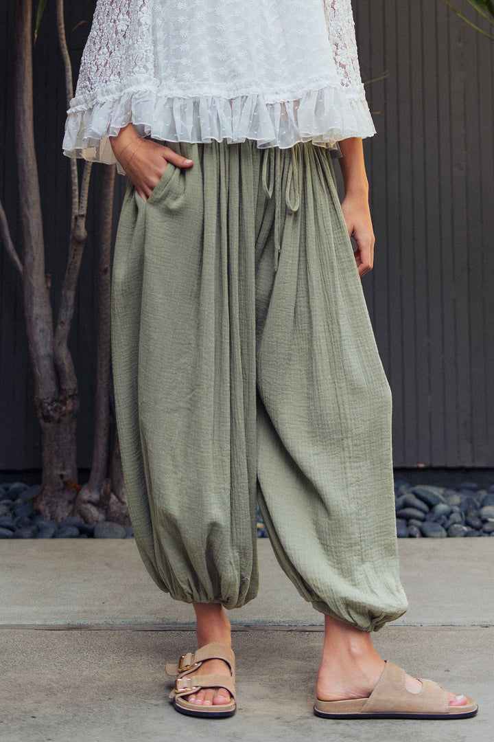 Woman wearing olive green crinkle cotton balloon pants with drawstring waist and elastic ankle cuffs paired with a white lace blouse.