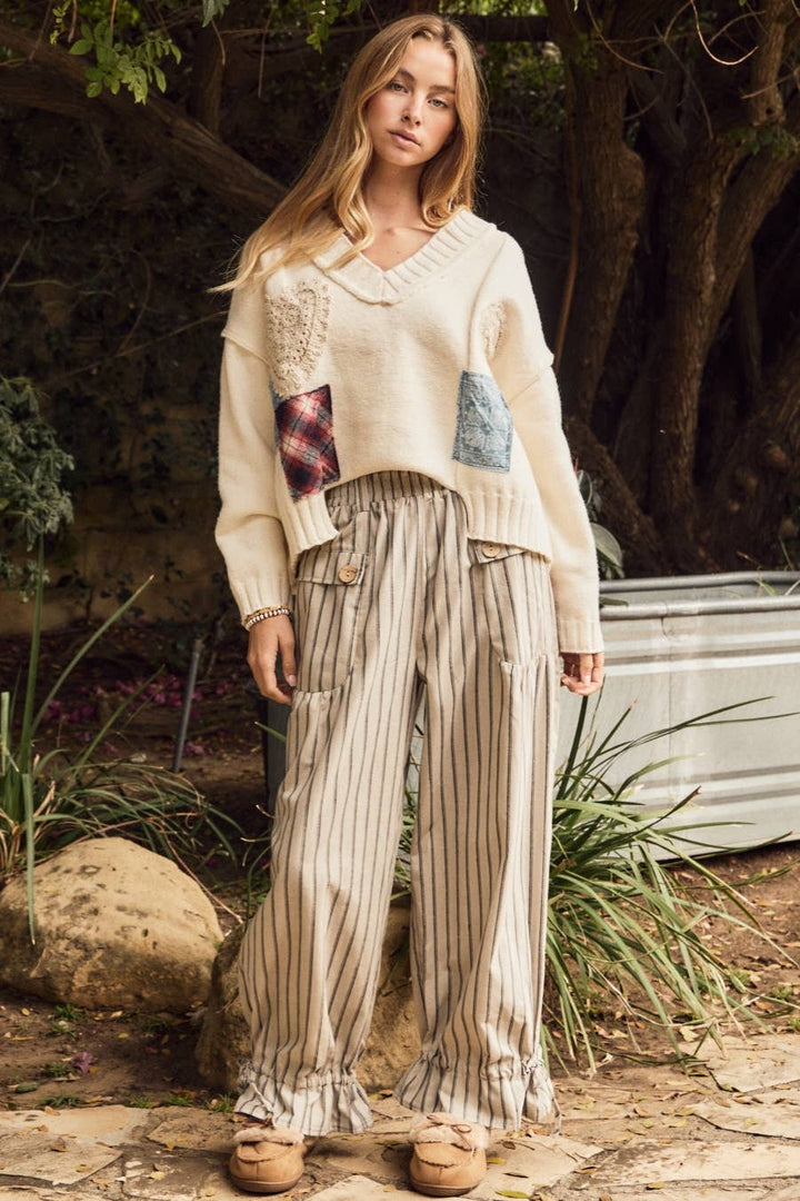 A woman stands outdoors wearing a cream knit sweater with patchwork details and wide-leg striped pants in beige and brown tones, paired with tan loafers, set against a natural background of trees and plants.