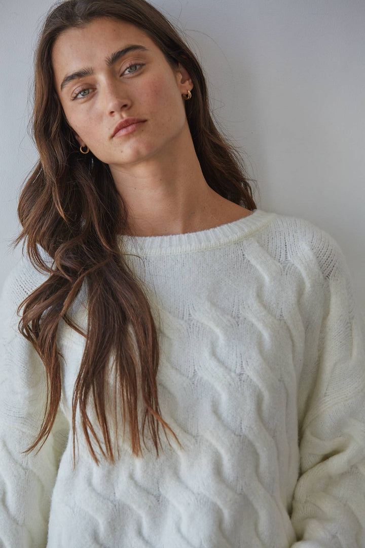 A woman with long wavy brown hair wearing a cream-colored cable-knit sweater, photographed against a plain light gray wall.