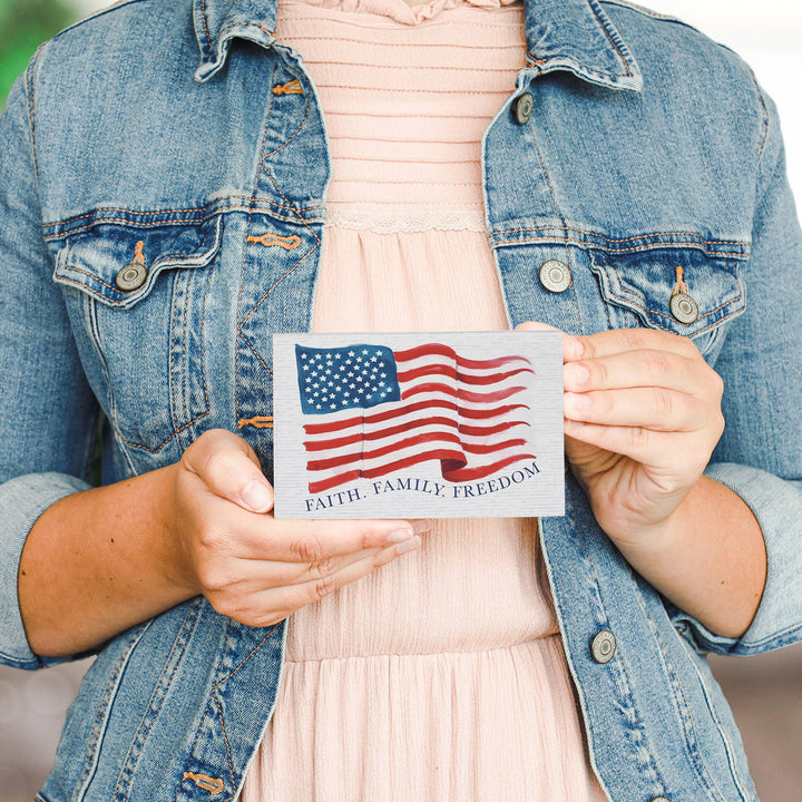 Person holding a wooden block with an American flag and text 'Faith. Family. Freedom' against a neutral background