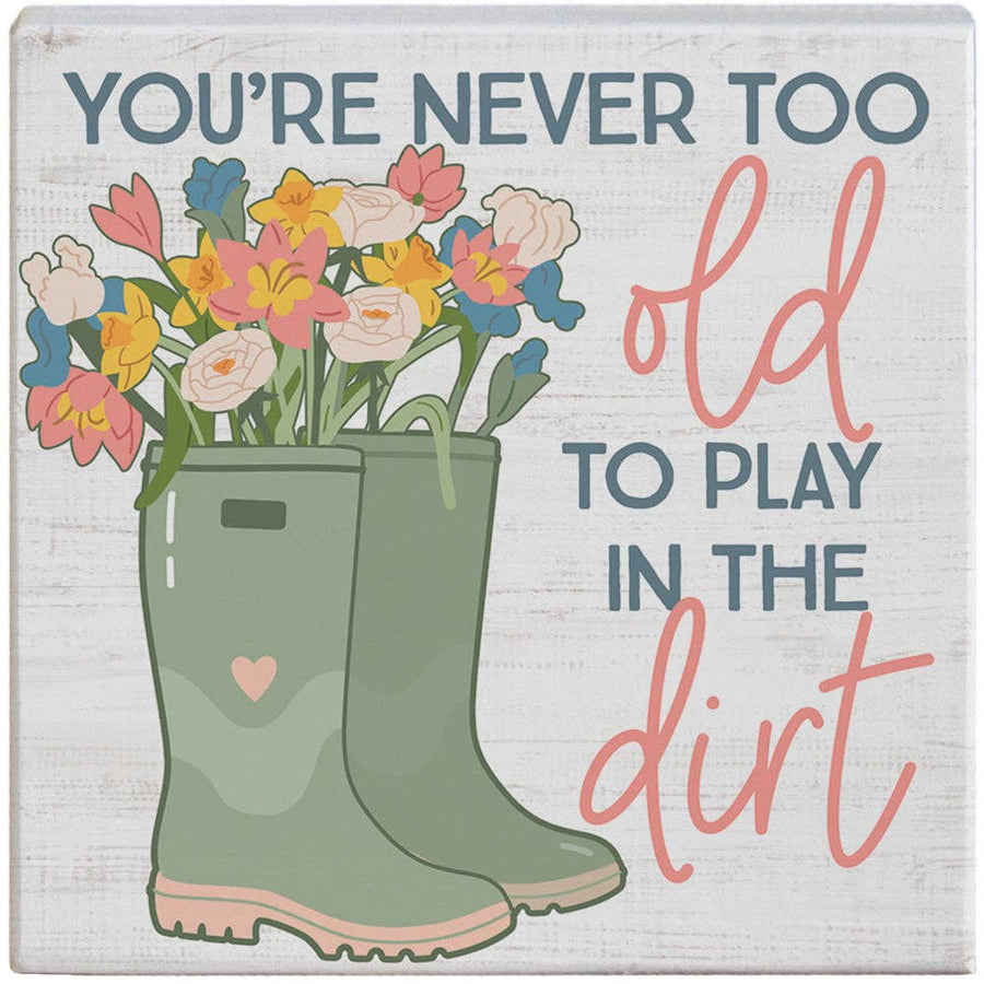 Decorative sign with green wellies, flowers, and text 'You're never too old to play in the dirt' on a light background.
