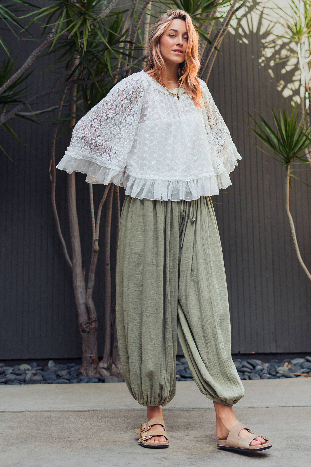Woman wearing olive green crinkle cotton balloon pants with drawstring waist and elastic ankle cuffs paired with a white lace blouse.