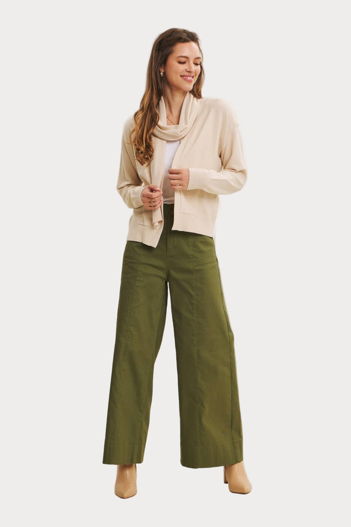 Woman wearing a beige cardigan, white shirt, and green pants on a plain background