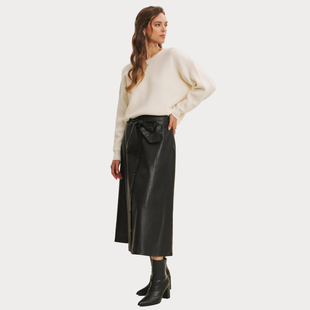 Woman wearing a Bow Back Sweater | Dolman Sleeve Knit with Satin Tie paired with a faux black leather skirt on a beige background. Side profile
