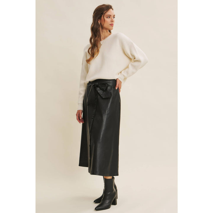 Woman wearing a Bow Back Sweater | Dolman Sleeve Knit with Satin Tie paired with a faux black leather skirt on a beige background. Side profile