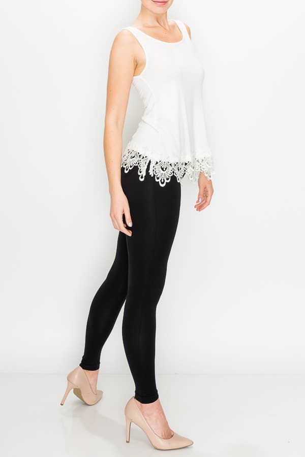 A model wears a sleeveless, scoop-neck tank top in ivory with a wide, intricate lace trim along the bottom hem. She is pairing it with black leggings and neutral-colored heels.

