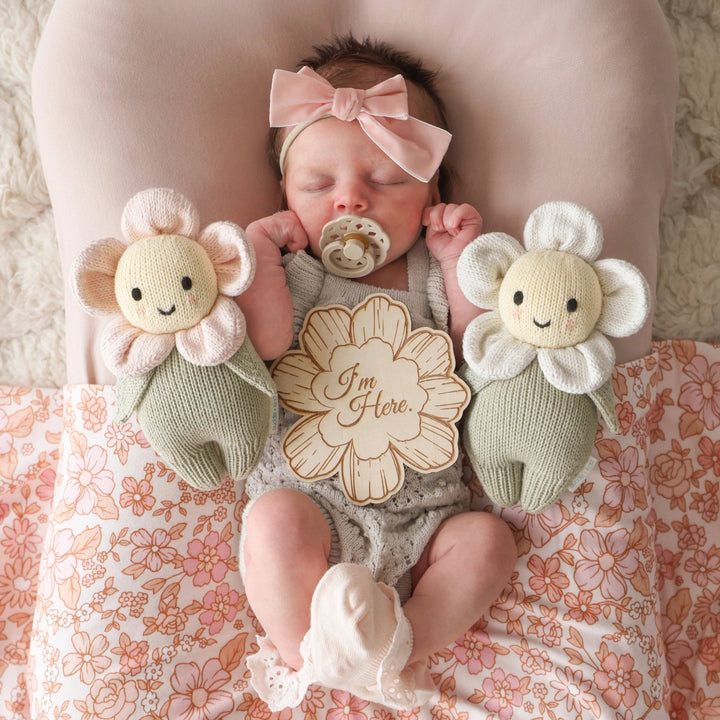 Baby lying on a floral blanket with flower-shaped toys and a 'I'm Here.' wooden card.