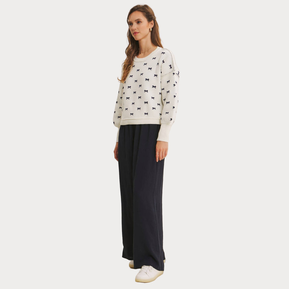 Model wearing an ivory jacquard knit sweater with a repeating navy blue bow pattern and voluminous balloon sleeves. Paired with wide-leg navy pants and white sneakers, styled for a casual, chic look.

