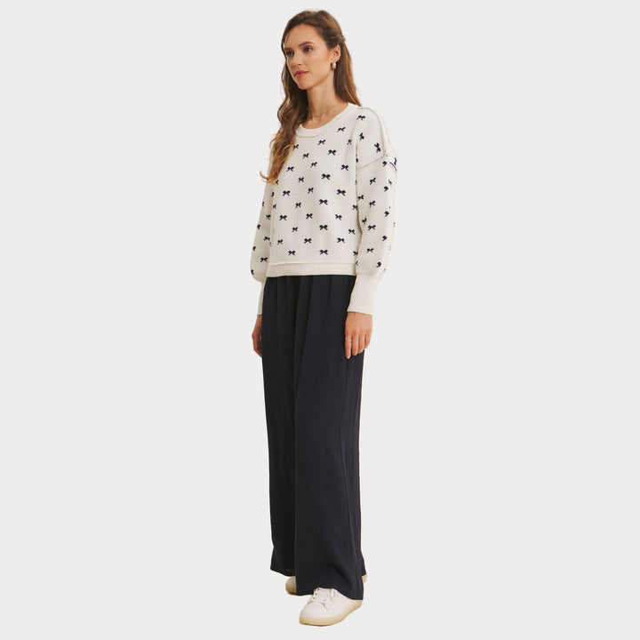 Model wearing an ivory jacquard knit sweater with a repeating navy blue bow pattern and voluminous balloon sleeves. Paired with wide-leg navy pants and white sneakers, styled for a casual, chic look.

