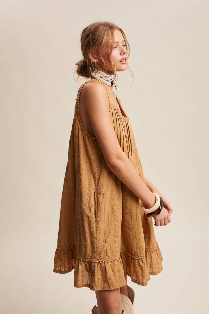 Woman wearing a textured camel-colored sleeveless mini dress with pintuck details and ruffle hem.