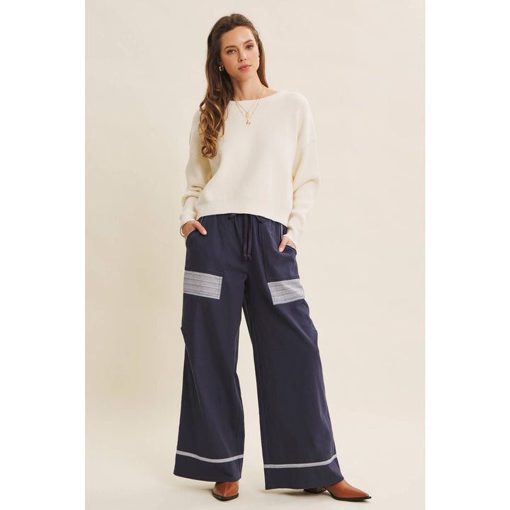 Model wearing Bow Back Sweater | Dolman Sleeve Knit with Satin Tie paired with navy pants with white lace patches and striped cuff on beige background