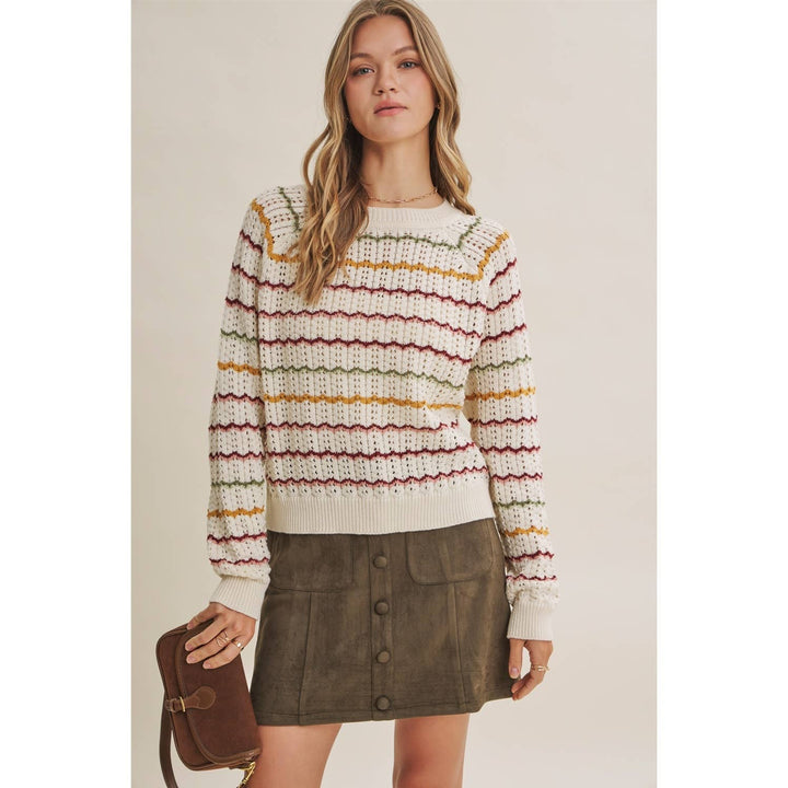 A woman models a cream knit sweater with horizontal stripes in rust red, mustard yellow, and olive green, paired with a brown mini skirt and holding a brown leather crossbody bag against a plain beige background.