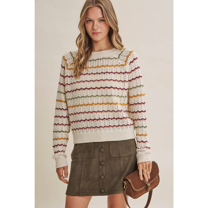 A woman models a cream knit sweater with horizontal stripes in mustard yellow, burgundy, and olive green, paired with an olive green mini skirt and a brown leather shoulder bag against a solid beige background.